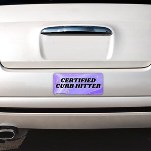 May include: A rectangular bumper sticker with a purple and white swirled background featuring the black text CERTIFIED CURB HITTER and small yellow star icons. The sticker is applied to the rear bumper of a white car with red taillights.