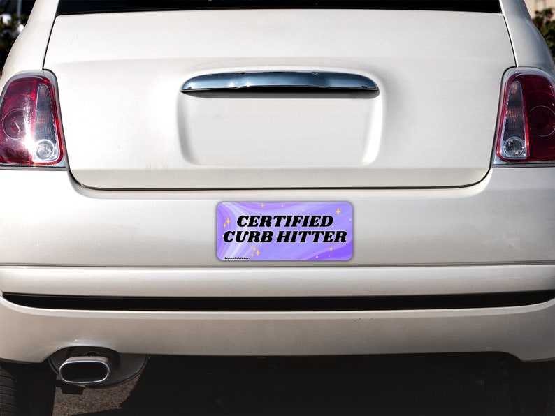 May include: A rectangular bumper sticker with a purple and white swirled background featuring the black text CERTIFIED CURB HITTER and small yellow star icons. The sticker is applied to the rear bumper of a white car with red taillights.
