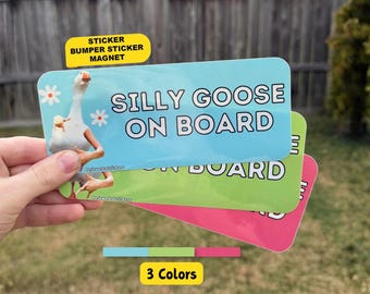 Może przedstawiać: Prostokątna naklejka lub magnes przedstawiający białego gęś z ludzkimi ramionami, białymi stokrotkami i napisem SILLY GOOSE ON BOARD. Produkt jest dostępny w kolorach jasnoniebieskim, limonkowym i jaskraworóżowym.