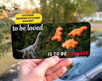 May include: A rectangular bumper sticker featuring a dark forest background with the white text to be loved and the text IS TO BE CHANGED in white and red. It displays a gray long-necked dinosaur and three orange dinosaur-shaped snacks.