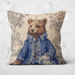 May include: A decorative throw pillow with a brown bear wearing a blue jacket and holding a teapot. The background features a floral pattern in shades of blue and white.
