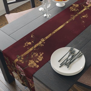 Peut inclure: Un chemin de table bordeaux avec une bande dorée et un motif floral. Le chemin de table est sur une table en bois foncé avec un bol de pommes, des verres à vin et des couverts.