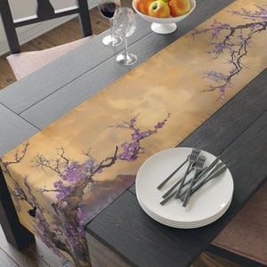 May include: A table runner with a floral design featuring a branch of pink blossoms against a yellow and brown background. The runner is laid on a dark wood table with two wine glasses, a bowl of apples, and a stack of white plates with silverware.