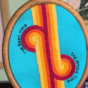 Op de afbeelding: Een ovale houten borduurring met turquoise stof voorzien van geborduurde verticale strepen in geel, oranje en donkerrood. Het borduurwerk bevat de zwarte tekst "I've Been Here" aan de linkerkant en "The Whole Time" aan de rechterkant.