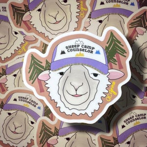 May include: A die-cut sticker featuring a white sheep wearing a purple and white baseball cap with the text "sheep camp counselor" printed on the front. The design includes small mountain icons and green pine trees against a light peach background.