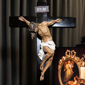 May include: A detailed sculpture of Jesus on a black wooden cross. The figure is depicted with realistic anatomy, wounds, and a white loincloth. The cross has a small sign at the top that reads "IN RI".