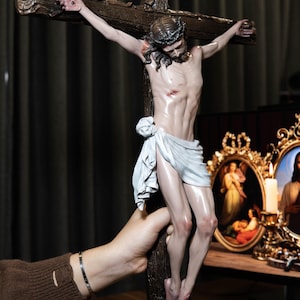 May include: A detailed sculpture of Jesus on a wooden cross. The figure is depicted with a crown of thorns and a white loincloth. The cross has the inscription "INRI" at the top. The sculpture is held by a hand.