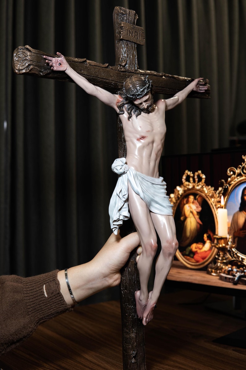 May include: A detailed sculpture of Jesus on a wooden cross. The figure is depicted with a crown of thorns and a white loincloth. The cross has the inscription "INRI" at the top. The sculpture is held by a hand.