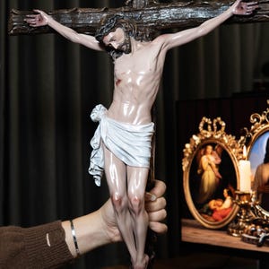 May include: A detailed sculpture of Jesus on a cross. The figure is depicted with a crown of thorns, a white loincloth, and a realistic depiction of the body. The cross is made of dark wood. The sculpture is held by a person.