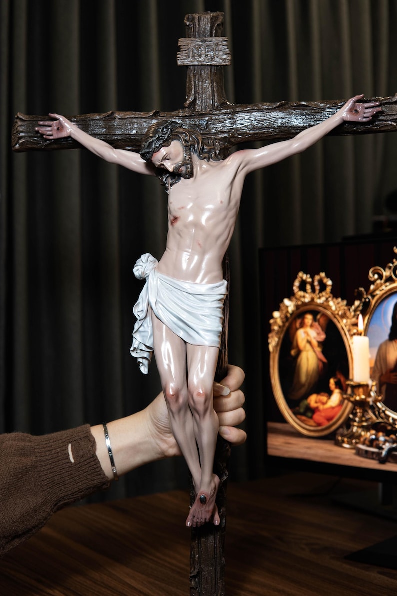 May include: A detailed sculpture of Jesus on a cross. The figure is depicted with a crown of thorns, a white loincloth, and a realistic depiction of the body. The cross is made of dark wood. The sculpture is held by a person.