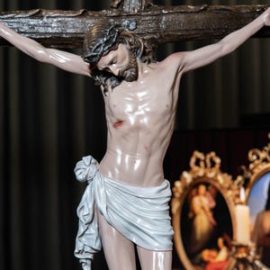 May include: A detailed sculpture of Jesus Christ on a wooden cross. The figure has a crown of thorns, a white cloth draped around the waist, and visible wounds. The background includes a dark curtain and framed artwork.