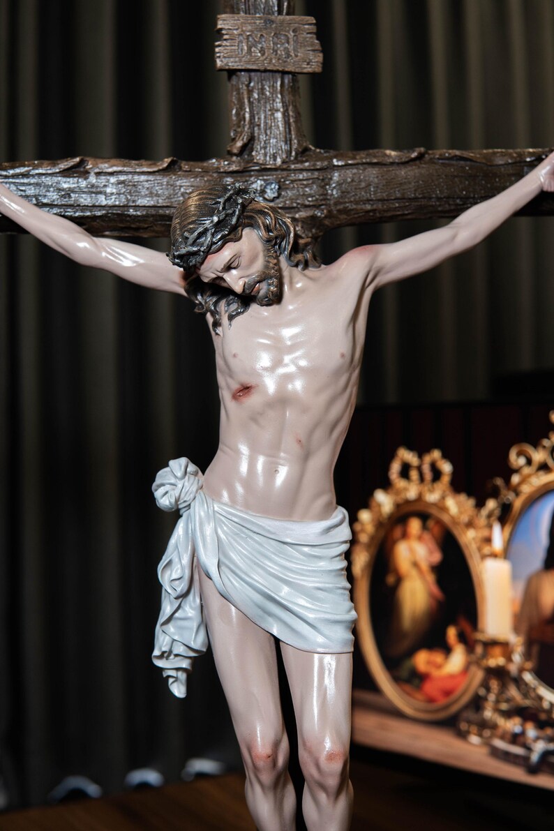 May include: A detailed sculpture of Jesus Christ on a wooden cross. The figure has a crown of thorns, a white cloth draped around the waist, and visible wounds. The background includes a dark curtain and framed artwork.
