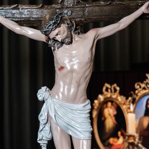 May include: A detailed statue of Jesus Christ on a wooden cross. The figure is depicted with a crown of thorns, a white cloth around the waist, and visible wounds. The background includes framed religious art.