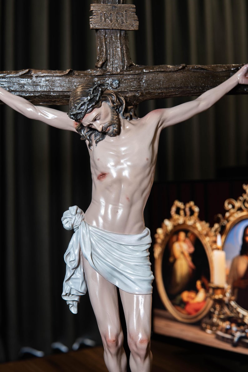 May include: A detailed statue of Jesus Christ on a wooden cross. The figure is depicted with a crown of thorns, a white cloth around the waist, and visible wounds. The background includes framed religious art.