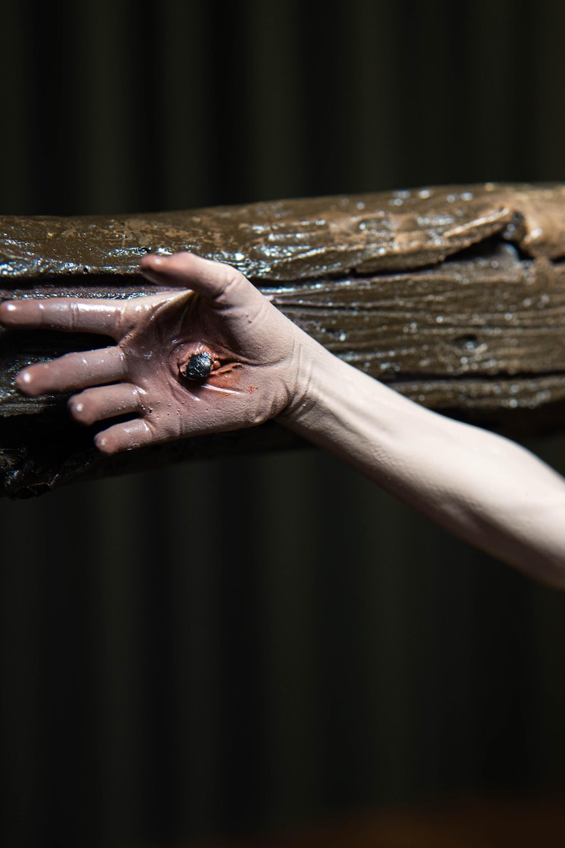 May include: A close-up of a hand with a dark wound, pierced by a dark object, possibly a nail. The hand is positioned against a rough, brown wooden beam. The background is dark.