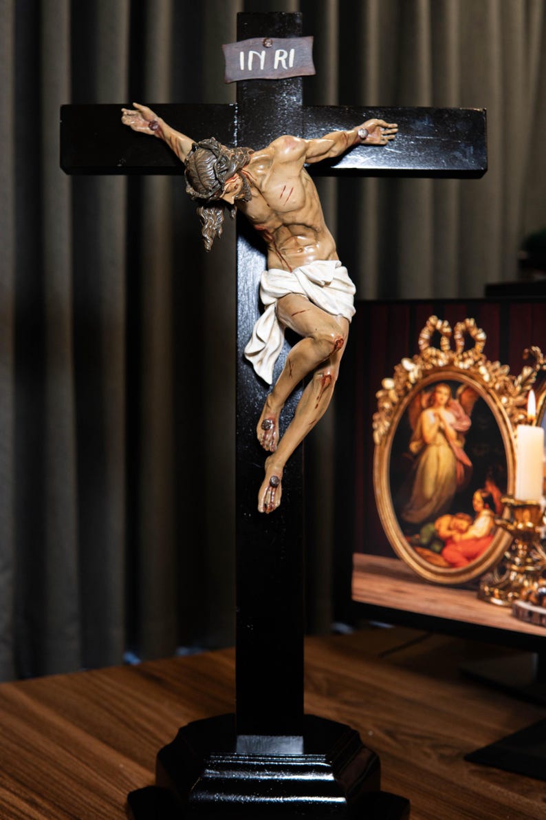 May include: A detailed sculpture of Jesus on a black wooden cross. The figure is depicted with realistic anatomy, wounds, and a white loincloth. The cross has a small sign at the top that reads "IN RI".
