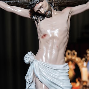 May include: A detailed statue of Jesus Christ on the cross. The figure is depicted with a crown of thorns, a beard, and a white cloth draped around the waist. The sculpture is painted with realistic skin tones and is mounted on a dark wooden cross.