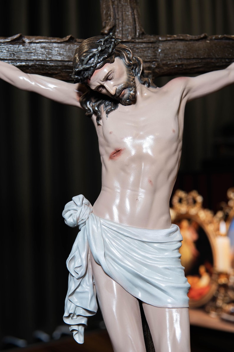 May include: A detailed statue of Jesus Christ on the cross. The figure is depicted with a crown of thorns, a beard, and a white cloth draped around the waist. The sculpture is painted with realistic skin tones and is mounted on a dark wooden cross.