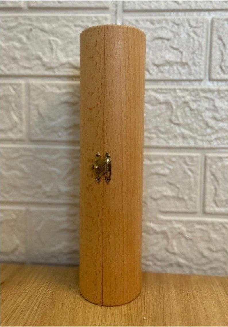 Puede incluir: Una caja cil&iacute;ndrica de madera con tapa abatible y cierre de lat&oacute;n. La caja es de color marr&oacute;n claro y tiene un acabado liso.