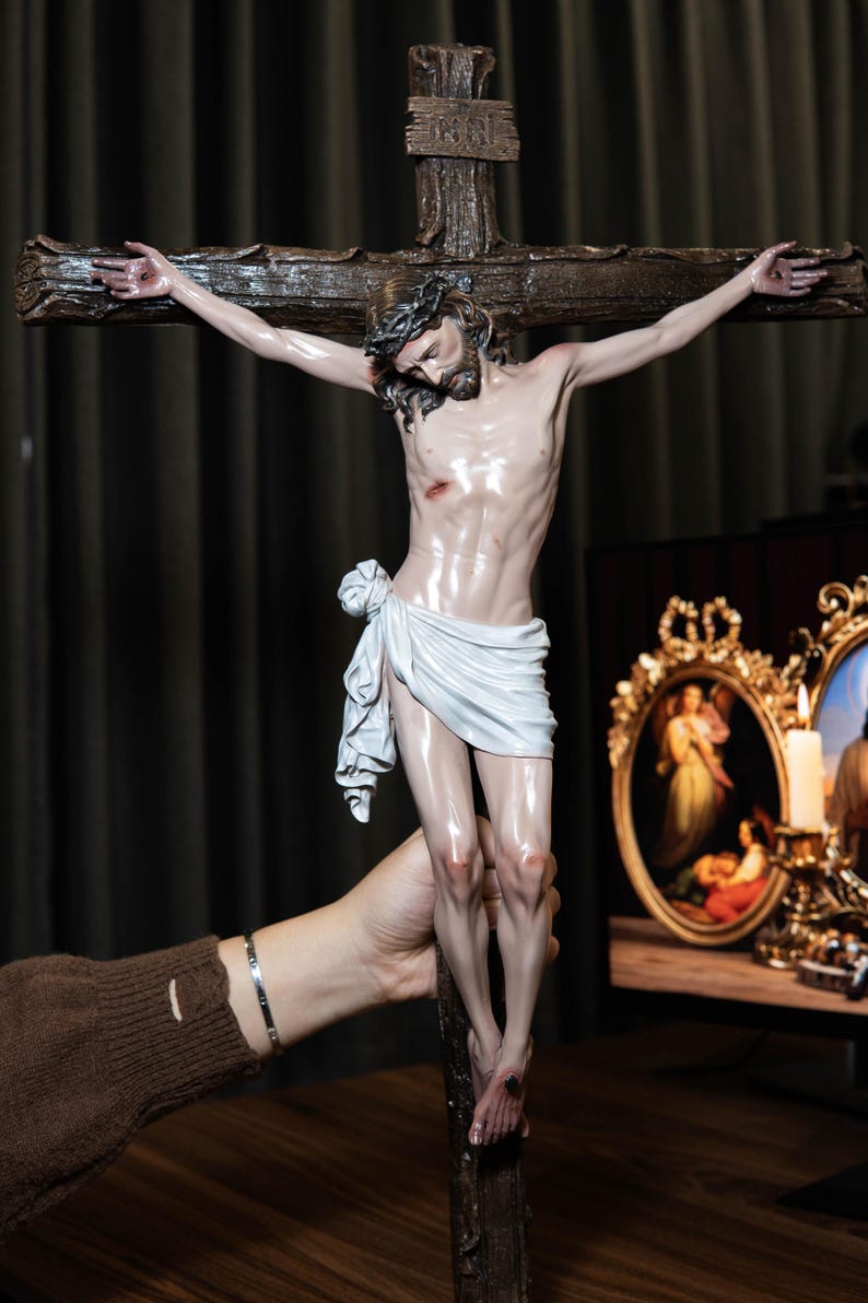 May include: A detailed sculpture of Jesus on a cross. The figure is depicted with a crown of thorns, a loincloth, and wounds. The cross is made of dark wood. The sculpture is held by a person.