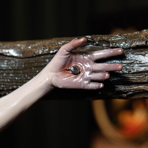 May include: A close-up of a hand with a nail through the palm, set against a dark background. The hand is pale with a reddish hue around the wound. The nail is dark and metallic, and the wood is textured and brown.