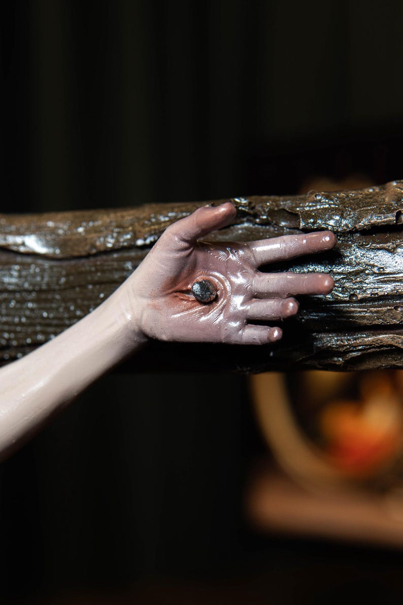 May include: A close-up of a hand with a nail through the palm, set against a dark background. The hand is pale with a reddish hue around the wound. The nail is dark and metallic, and the wood is textured and brown.