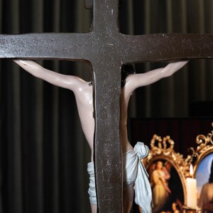 May include: A detailed view of a crucifix with a dark, textured cross. The figure of Jesus is depicted with a pale complexion and a white cloth around the waist. The background is a dark curtain.