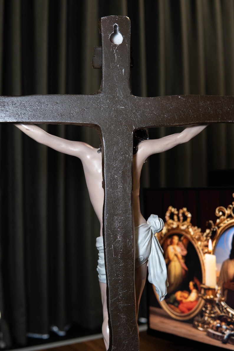 May include: A detailed view of a crucifix with a dark, textured cross. The figure of Jesus is depicted with a pale complexion and a white cloth around the waist. The background is a dark curtain.