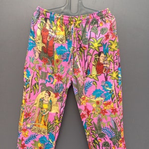 May include: Pink pajama pants with an all-over floral and figure print. The pants feature a drawstring waist and white trim at the cuffs. The print includes images of people, flowers, and foliage in shades of red, yellow, blue, and green.