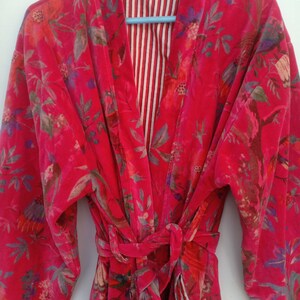 May include: A vibrant red velvet robe with a floral pattern in shades of pink, purple, and green. The robe has long sleeves and a matching belt. The interior lining features a striped pattern in red and white.