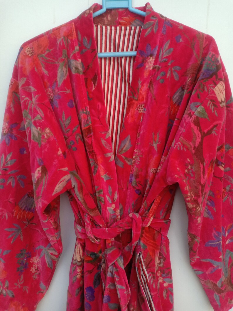May include: A vibrant red velvet robe with a floral pattern in shades of pink, purple, and green. The robe has long sleeves and a matching belt. The interior lining features a striped pattern in red and white.
