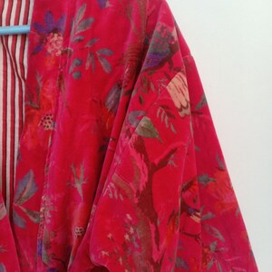May include: A vibrant red velvet robe with a floral pattern in shades of pink, purple, and green. The robe's interior lining features a striped pattern in red and white. The robe is hanging on a blue hanger.