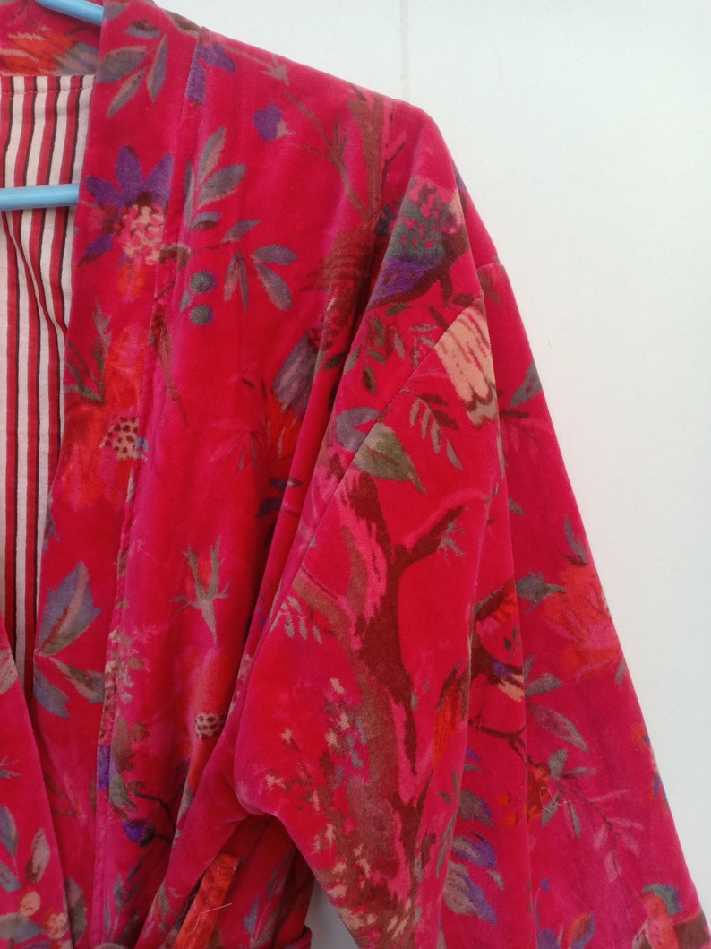 May include: A vibrant red velvet robe with a floral pattern in shades of pink, purple, and green. The robe's interior lining features a striped pattern in red and white. The robe is hanging on a blue hanger.