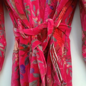 May include: A vibrant pink floral robe with a matching belt. The robe features a colorful pattern of flowers and leaves in shades of red, purple, and gray. The robe has a pocket with a striped lining.