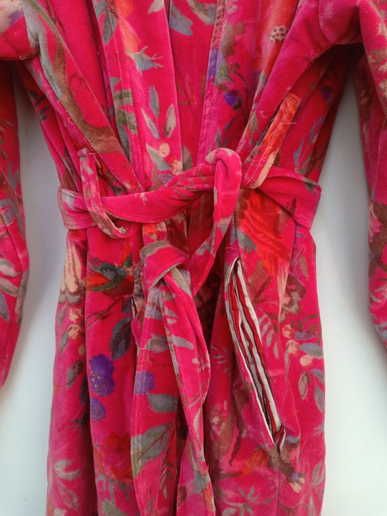 May include: A vibrant pink floral robe with a matching belt. The robe features a colorful pattern of flowers and leaves in shades of red, purple, and gray. The robe has a pocket with a striped lining.