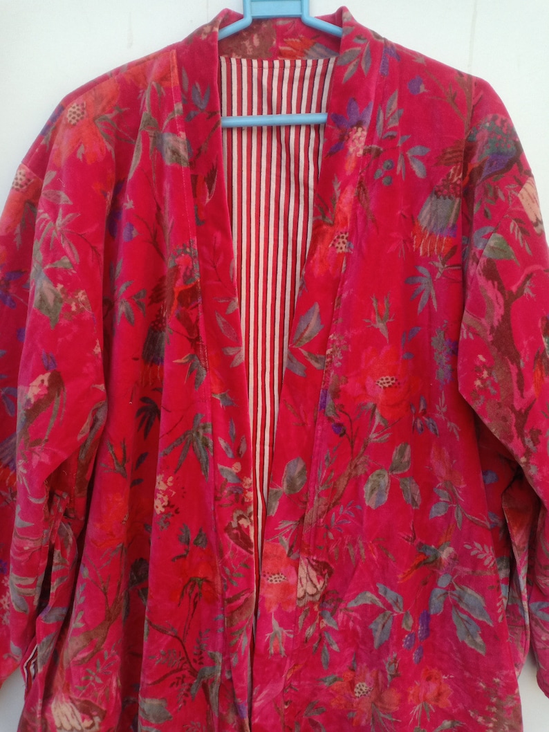 May include: A vibrant red floral print kimono-style jacket. The jacket features a bold pattern of flowers and foliage in shades of pink, green, and purple. The interior lining has a white and red striped pattern. The jacket is hanging on a blue hanger.