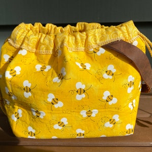 May include: A yellow drawstring bag with a brown leather handle. The bag is decorated with a pattern of white bees on a yellow background.