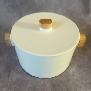 Puede incluir: Olla de cerámica blanca con tapa de madera y asas de madera. La olla es redonda y tiene una superficie lisa.