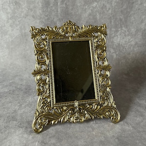 May include: An ornate picture frame in a gold tone, featuring a decorative, scrolled design. The frame has a rectangular mirror inside.