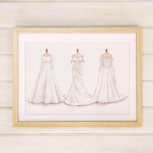 May include: A framed print featuring three white wedding dress designs. The dresses are displayed on mannequins, showcasing different styles: long-sleeved, strapless, and off-the-shoulder. The frame is a light wood color, set against a white wooden background.
