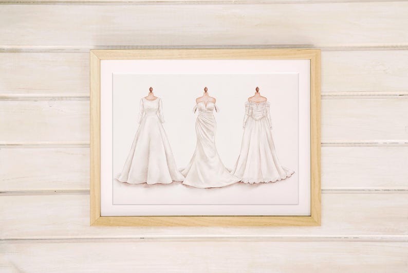 May include: A framed print featuring three white wedding dress designs. The dresses are displayed on mannequins, showcasing different styles: long-sleeved, strapless, and off-the-shoulder. The frame is a light wood color, set against a white wooden background.
