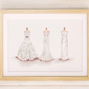 May include: A framed print featuring three white wedding dresses on dress forms. The dresses vary in style, with one having a full skirt, one a mermaid silhouette, and the third a simple sheath design. The frame is light wood.