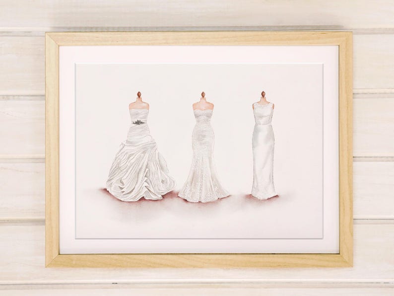 May include: A framed print featuring three white wedding dresses on dress forms. The dresses vary in style, with one having a full skirt, one a mermaid silhouette, and the third a simple sheath design. The frame is light wood.
