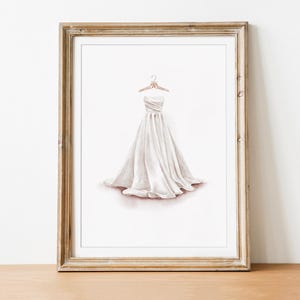 May include: Watercolor print of a white strapless wedding dress on a hanger. The dress has a flowing skirt and is framed in a gold-toned wooden frame. The artwork is displayed on a wooden surface against a white wall.