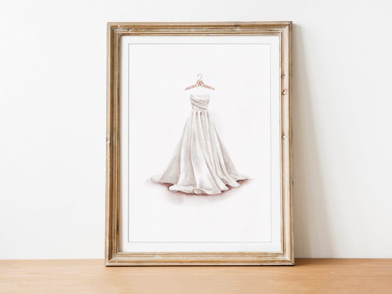 May include: Watercolor print of a white strapless wedding dress on a hanger. The dress has a flowing skirt and is framed in a gold-toned wooden frame. The artwork is displayed on a wooden surface against a white wall.