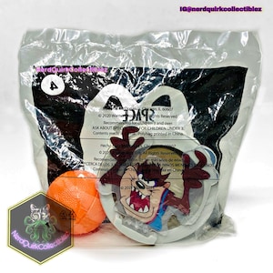 May include: A plastic bag containing a Happy Meal toy featuring a cartoon Tasmanian Devil holding a basketball. The bag is white with blue and red text that reads "© 2020 Warner Bros. Ent. All Rights Reserved. Recommended for children 3 and over. Ask about special toys for children under 3. Contents made in China. Polybag printed in China."