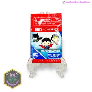 Puede incluir: Una bolsa de plástico roja y azul con una etiqueta de advertencia sobre el riesgo de asfixia. La bolsa contiene un paquete de pegatinas HeroBadges de DC Comics. El paquete de pegatinas presenta a Superman, Batman y Wonder Woman. La bolsa está etiquetada con "ONLY at CIRCLE K".