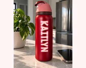 May include: A red Owala water bottle with a pink lid and a push-button mechanism. The name KAITLYN is printed vertically in large, light pink capital letters with a white outline on the side of the cylindrical metal bottle body.