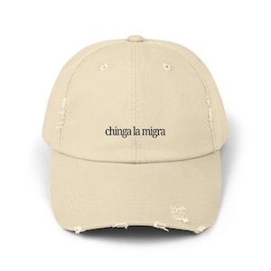 May include: A beige baseball cap with a distressed, worn-in finish featuring the black embroidered text chinga la migra centered on the front panel. The hat includes a curved brim, stitched eyelets, and a structured crown design.