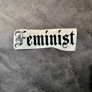 Może przedstawiać: Biała naklejka z napisem "Feminist" w czcionce gotyckiej. Naklejka jest prostokątna i ma lekko postrzępioną krawędź. Tekst jest wyśrodkowany na naklejce.